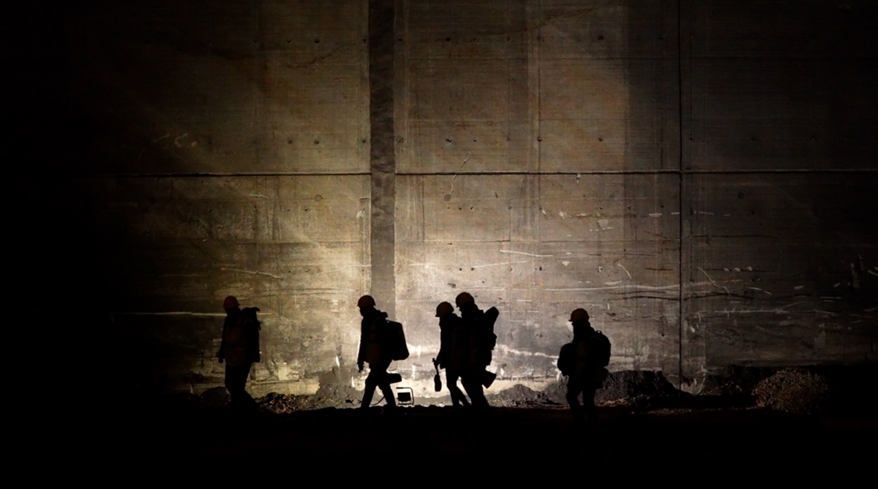 HOOU-Projekt WATTwanderungen: Moorburg-Film transformation Eine schattenhafte Gruppe von fünf Menschen mit Bauhelmen, dicken Jacken, Rucksäcken bewegt sich im Scheinwerferlicht an einer dunklen Mauer entlang